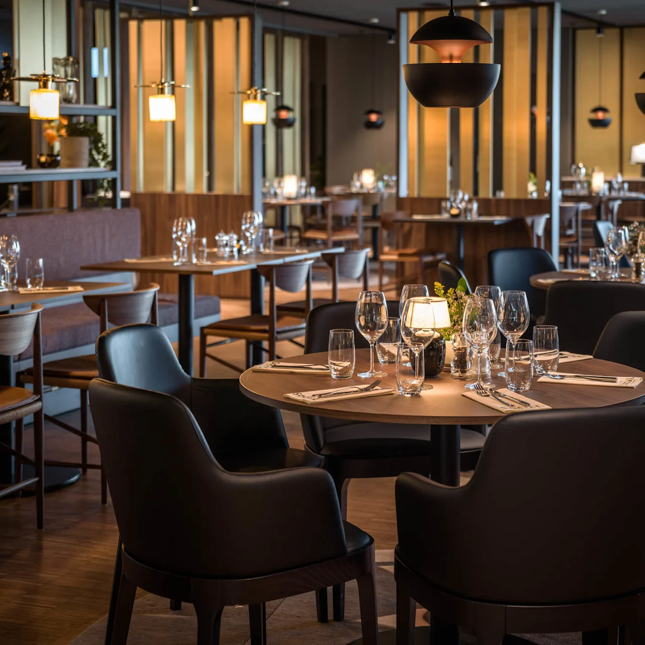 Et rundt bord dekket med vinglass og bestikk står under dempet belysning i en vakkert dekorert restaurant med polstrede stoler og trefordelinger. 