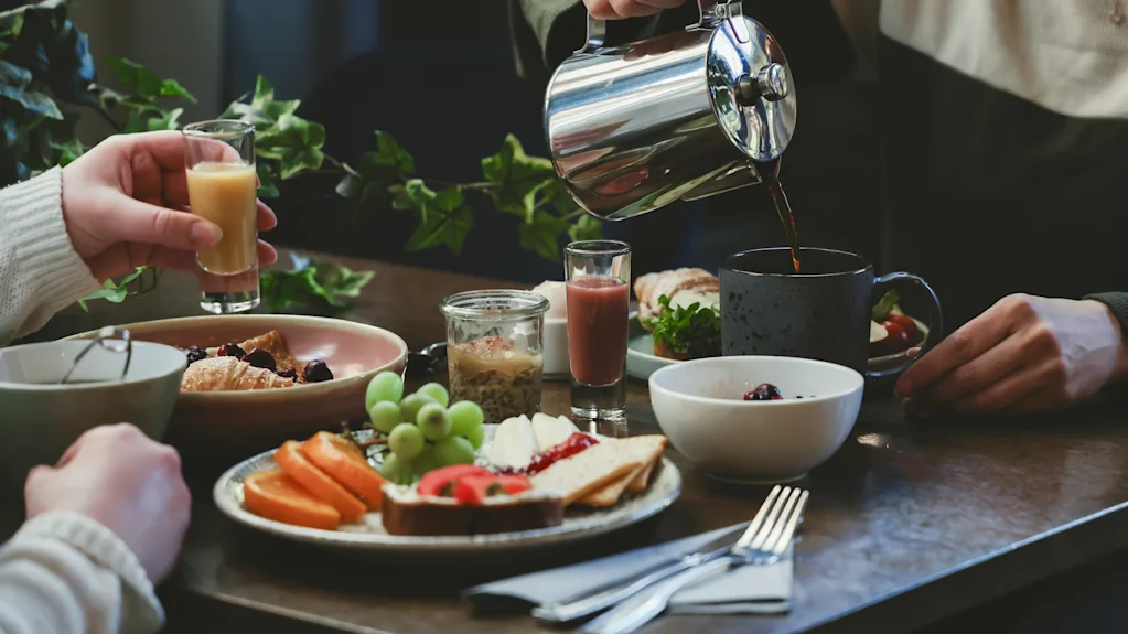 En person häller kaffe från en kanna i en mugg på ett bord med frukostmat inklusive frukt, bakverk och drycker, omgivet av gröna växter.