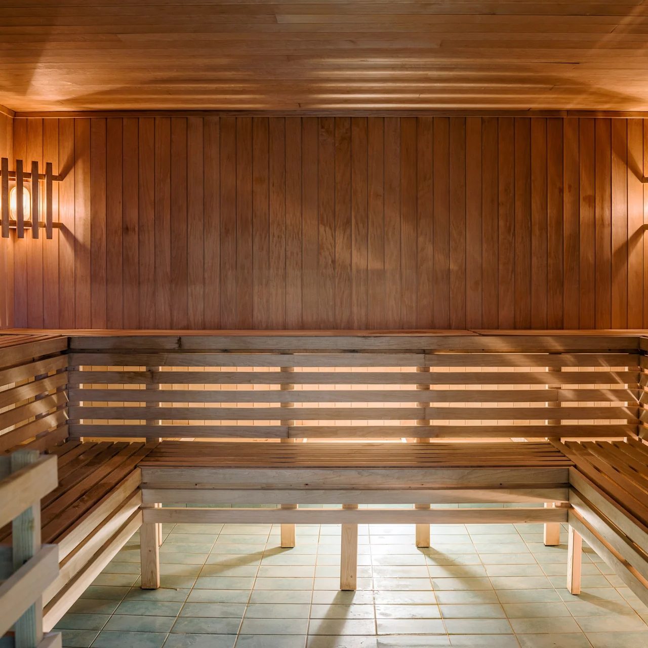 Wooden benches line the corners of a sauna, softly illuminated by warm, wall-mounted lights. The wood-paneled walls and ceiling create a cozy, enclosed atmosphere. No text present.