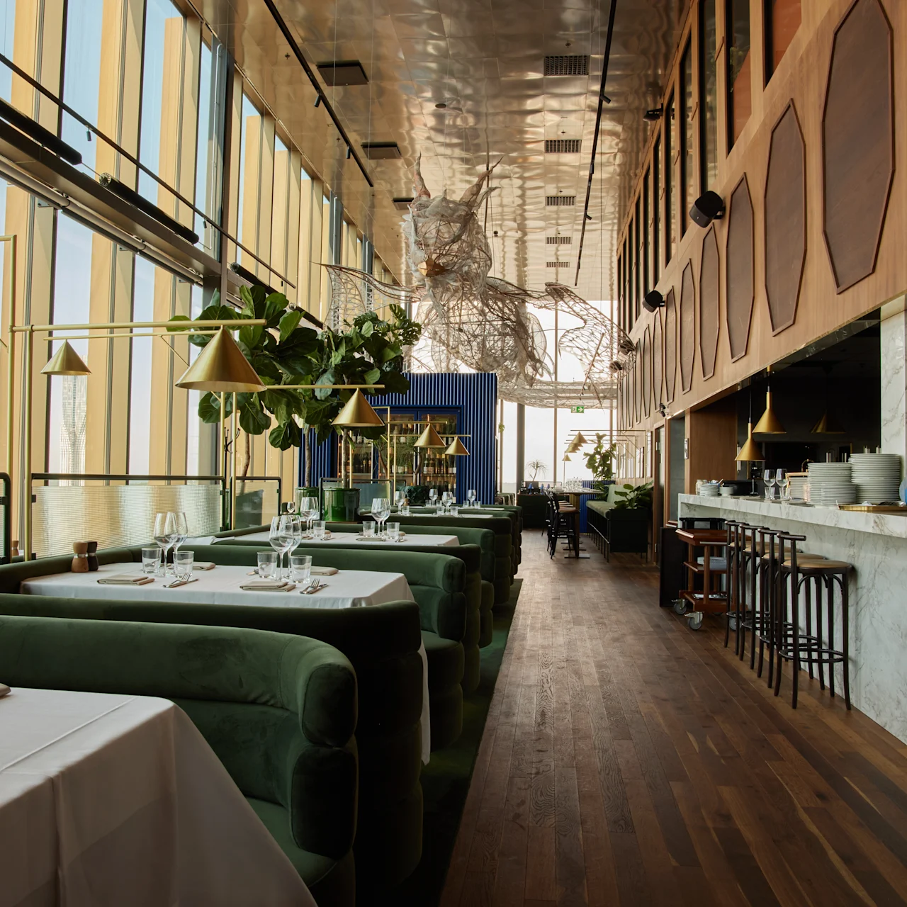 Bord och stolar är uppställda i en elegant restaurang, med gröna klädslar och en bardisk i marmor. Rummet är fyllt av naturligt ljus från stora fönster och dekorerat med hängande lampor.