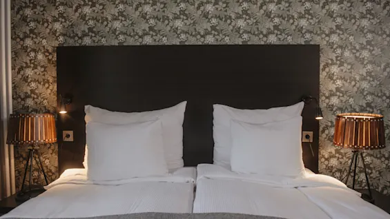 Bed with two large pillows, lit by bedside lamps on each side, in front of a floral wallpaper.