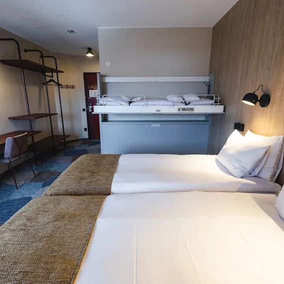Ett hotellrum med en dubbelsäng, prydligt bäddad, har en våningssäng på väggen. Moderna belysningsarmaturer framhäver ett skrivbord och en stol längs ett mönstrat golv.