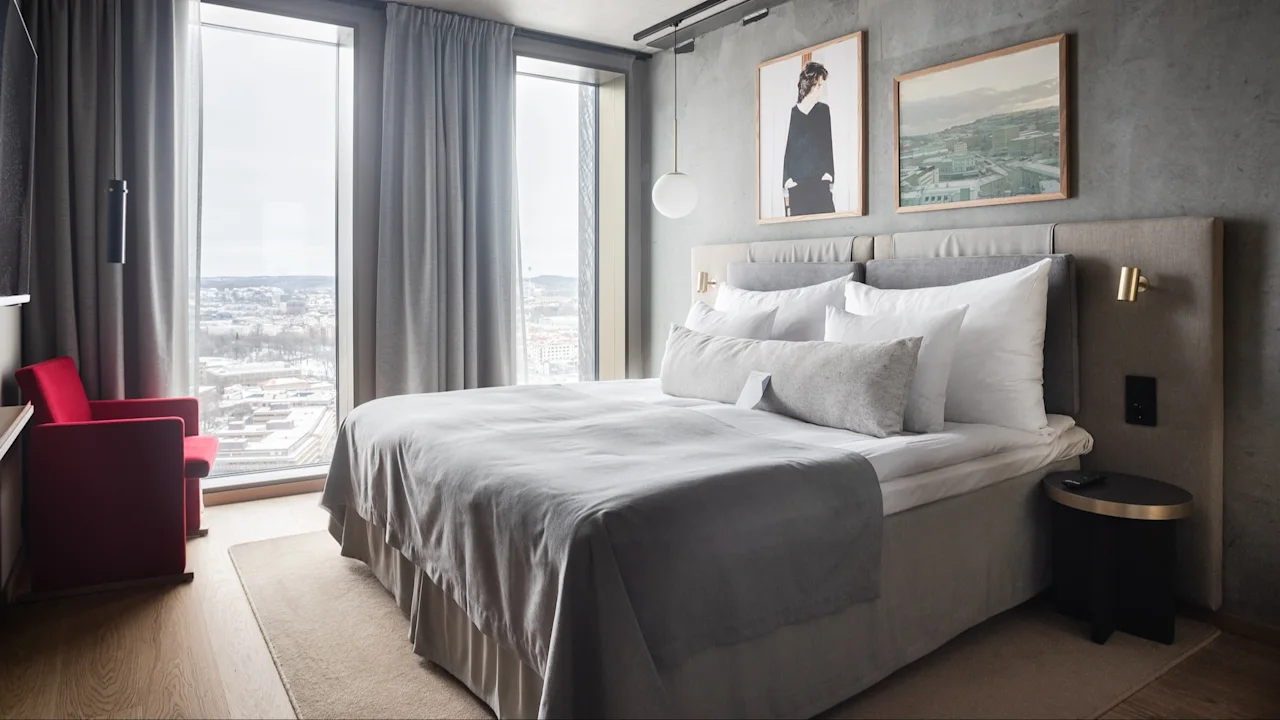 A neatly made bed with gray linens is set against a wall with framed art, beside floor-to-ceiling windows and a red chair. 