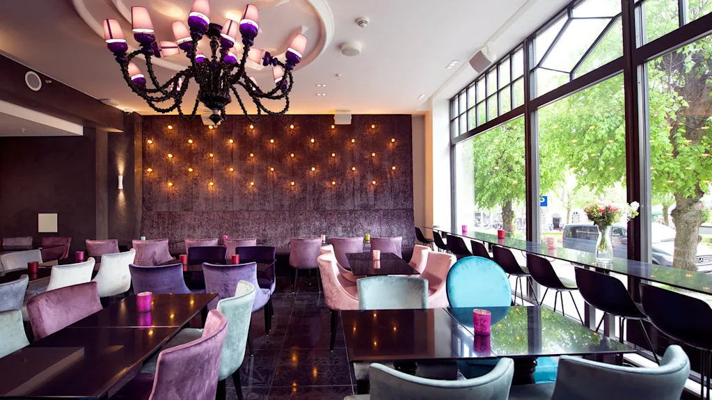 En stor, lilla lysekrone hænger over en moderne restaurant med farverige stole og mørke borde. Blød belysning dekorerer fjernevæggen, og store vinduer afslører træer udenfor.
