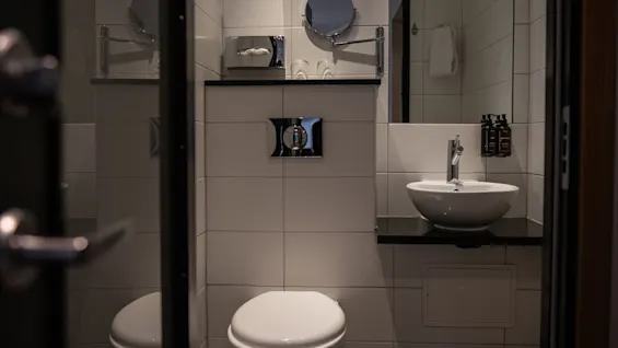 A well-lit, contemporary bathroom with white tiled walls, a toilet, a basin, and a mirror, suggesting comfort and cleanliness.