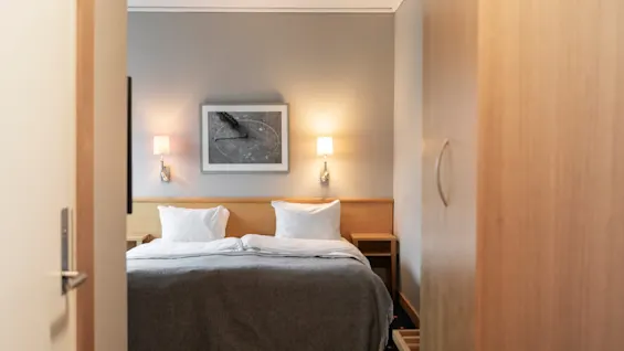 Et komfortabelt hotellrom med en seng, nattbord og vegglamper, sett gjennom en åpen døråpning.