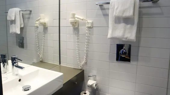 Et badeværelse viser en hvid flisevæg, et spejl, en hvid vask, en krom vandhane, en monteret hårtørrer og hvide håndklæder på en håndklædestang.