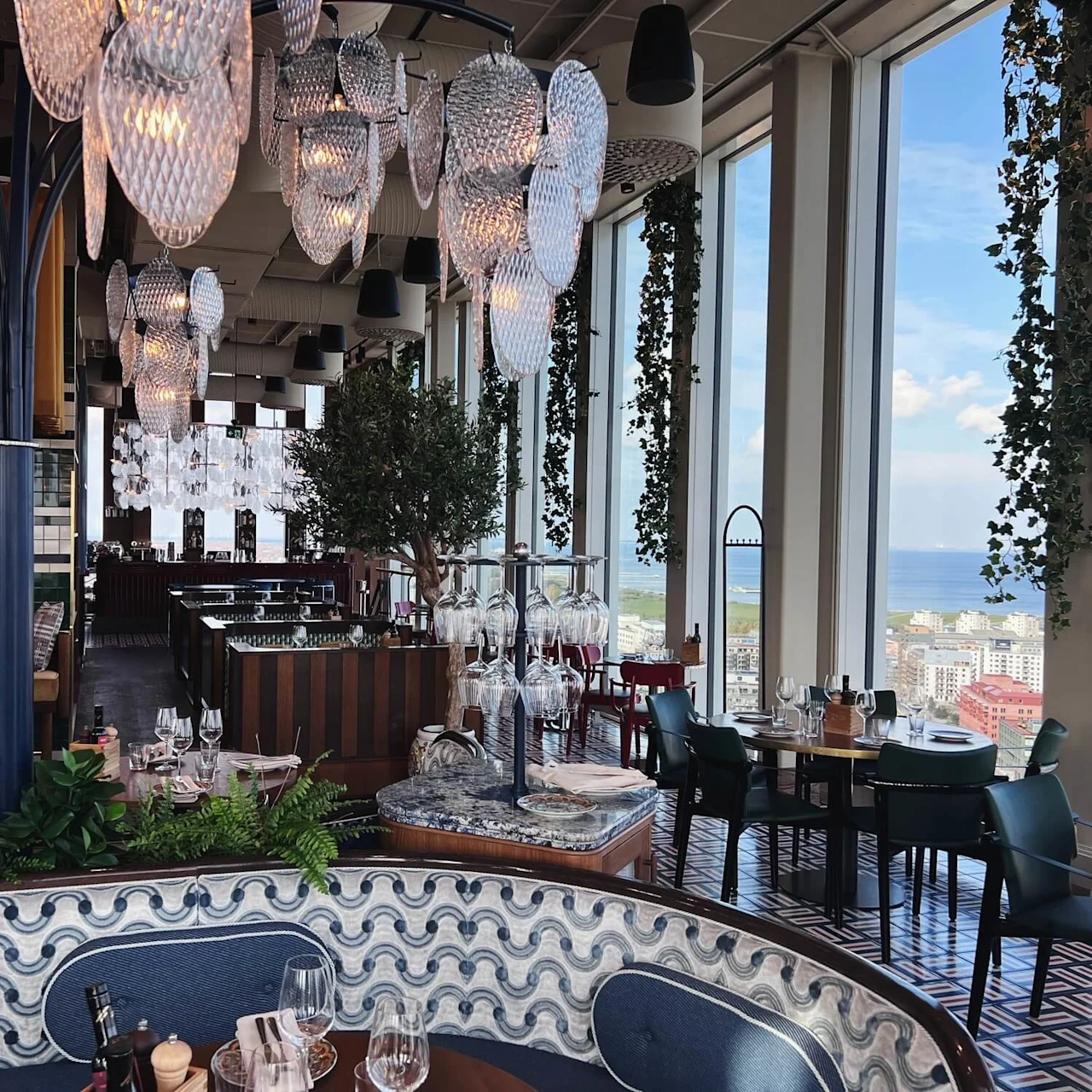 Luksuriøs restaurant med hængende glaskroner og frodig grønne områder. Spiseborde er elegant dækket tæt på store vinduer, med panoramaudsigt over by og hav. Elegant atmosfære med detaljeret indretning.