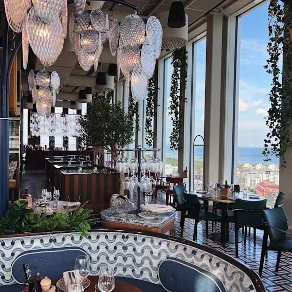 Lyxig restaurang med hängande glaskronor och frodig grönska. Matbord är elegant dukade nära stora fönster, med panoramautsikt över stad och hav. Elegant atmosfär med detaljerad inredning.
