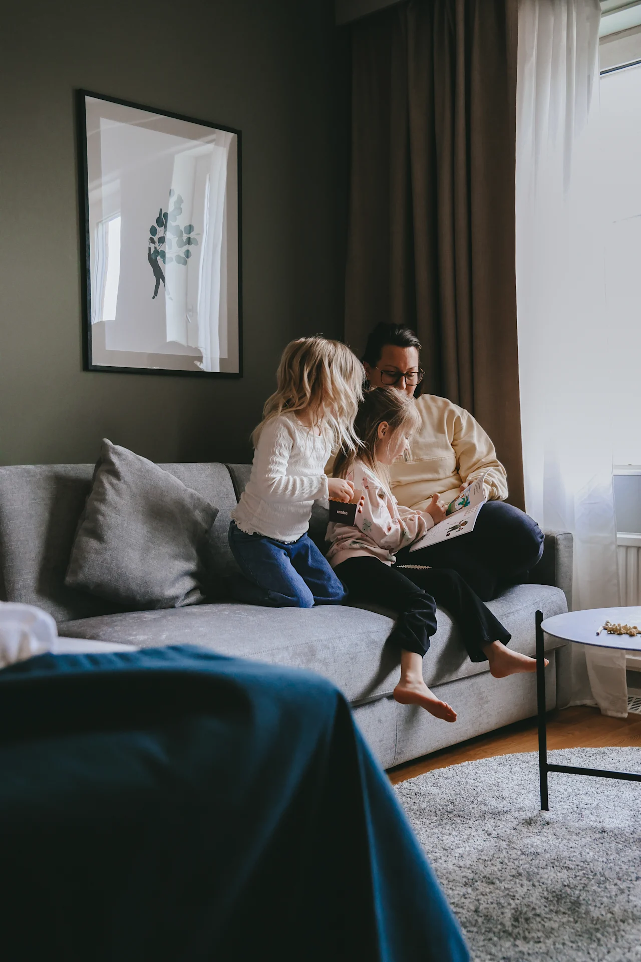 A family sits on a gray sofa, a parent reading a book to two children in a cozy living room with framed art and curtains.