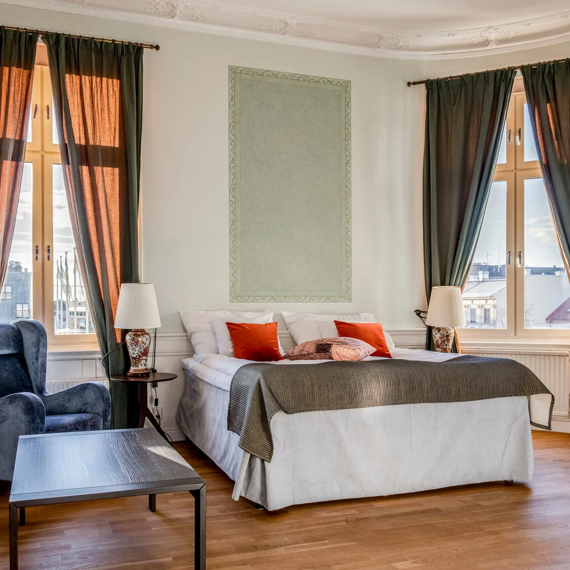 Elegantly furnished bedroom with a large bed, armchairs, and classic decor in a bright, spacious room with tall windows.