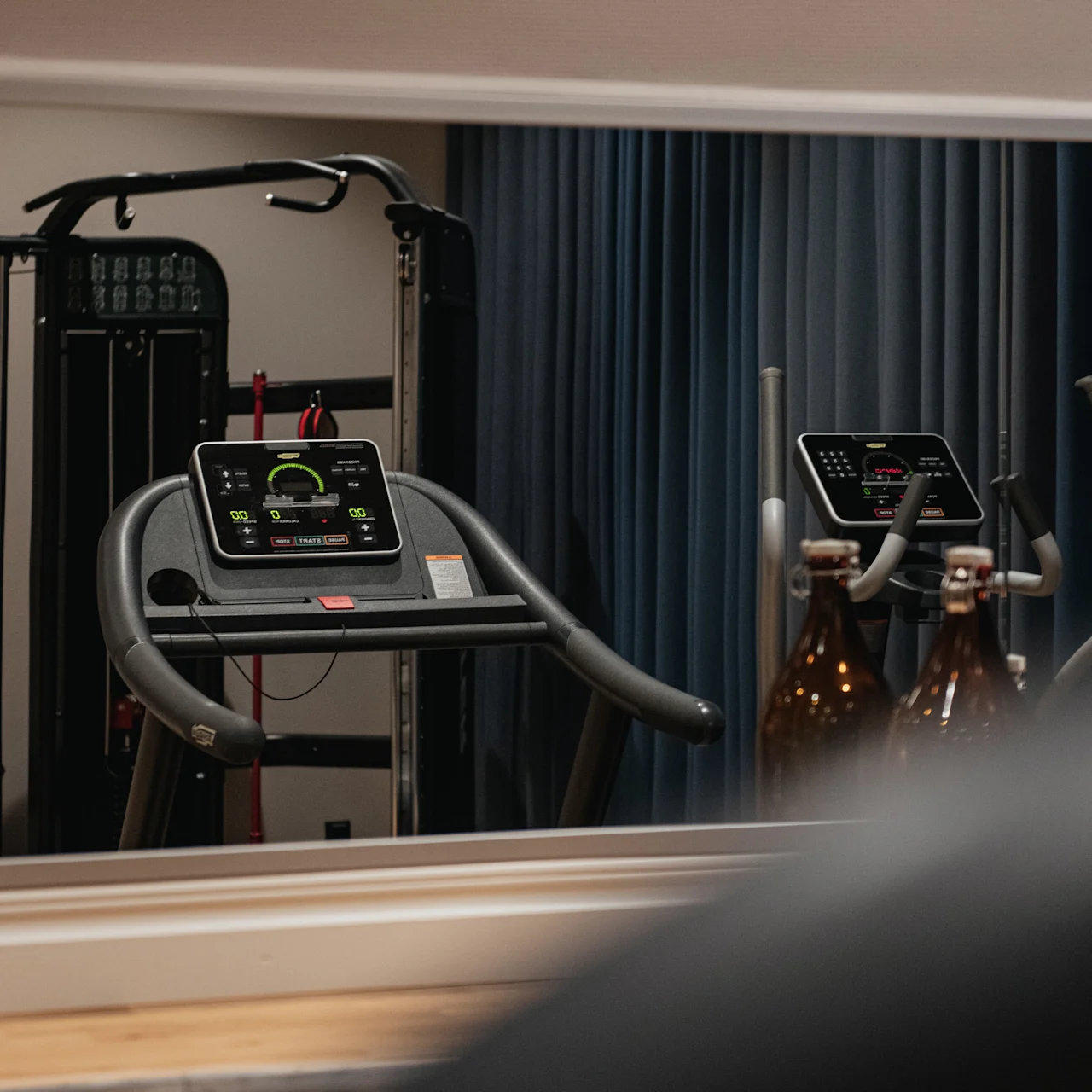 Löparbandsdisplay reflekteras i gymspegel; träningsutrustning runtom, blå gardiner i bakgrunden.