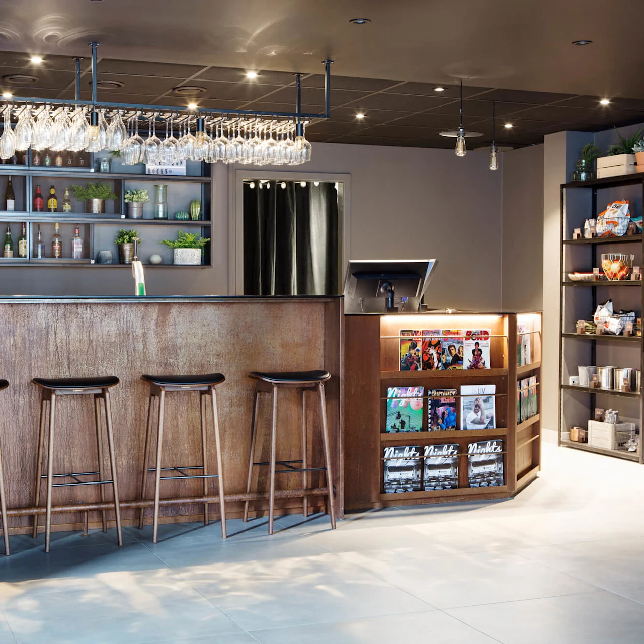 Bar med træstole står foran hylder med glas og flasker; i nærheden tilbyder en lille magasinholder læsemateriale i en varmt oplyst, moderne indretning.