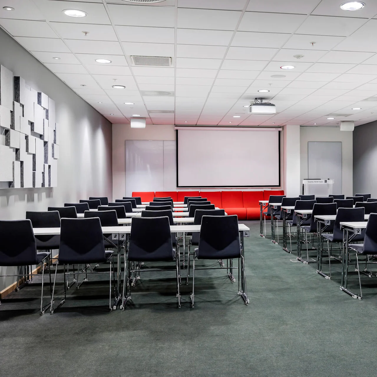 A conference room with rows of black chairs and tables faces a large projection screen. Red couches line the wall beneath the screen, surrounded by gray walls and acoustic panels.