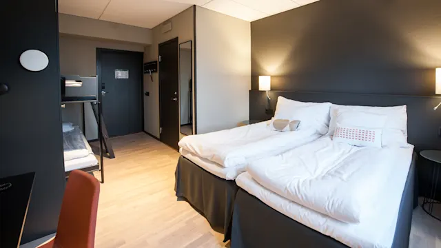 Siististi järjestetty hotellihuone, jossa on valkopedlinen parivuode kahden pienen seinälampun vieressä. Ovi ja naulakko näkyvät modernissa, siistissä tilassa.