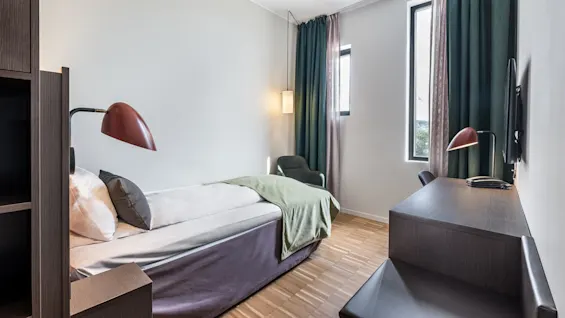 A single bed with a green blanket faces a small desk with a lamp and chair in a bright room with large windows and heavy curtains.