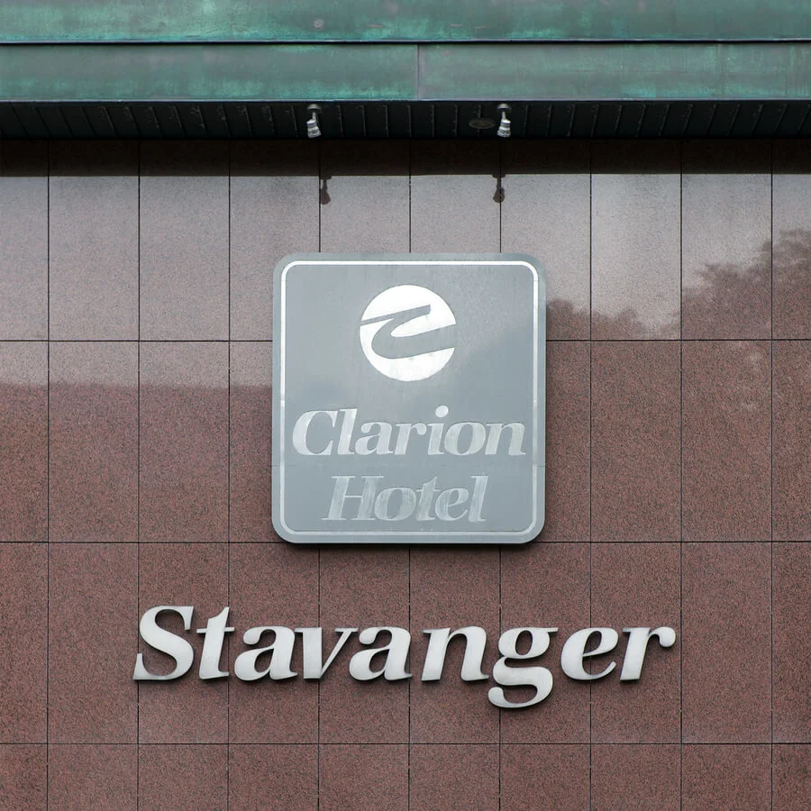 A sign displaying "Clarion Hotel Stavanger" is mounted on a red and gray tiled wall. Spotlights are fixed above, casting light downward. 