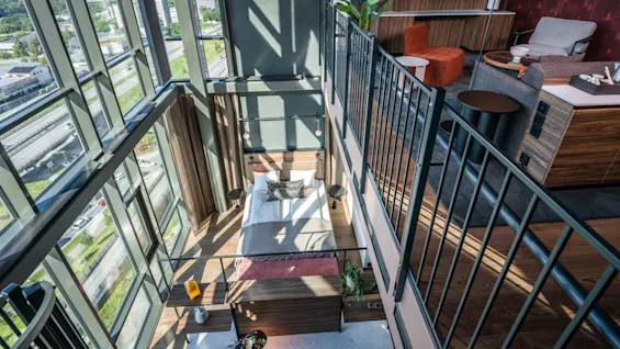Soverom under en mesanin med glassvegger som har utsikt over en by. Moderne møbler, seng, sofa, lenestol og en stor plante innredet i rommet.
