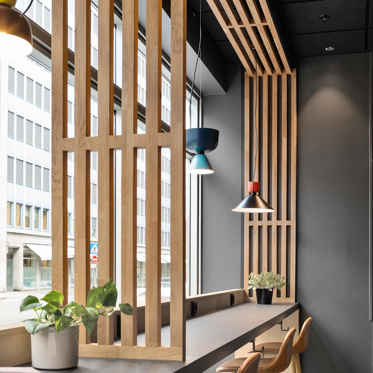 A stylish cafe interior features high stools along a wooden counter with potted plants and hanging lamps. Large windows overlook a city street, enhancing the bright, modern atmosphere.