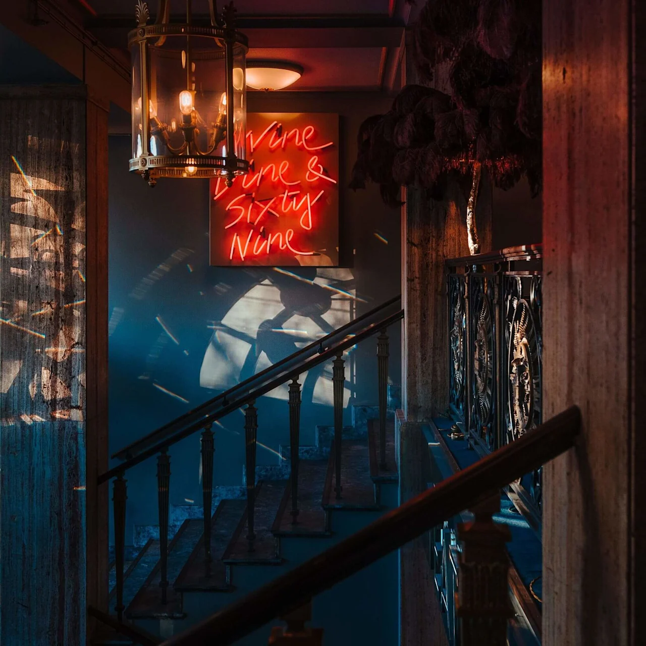 A warmly lit chandelier hangs in a dimly lit staircase. A neon sign on the wall reads "Wine Dine & Sixty Nine," casting colorful reflections on wooden railings and columned walls.