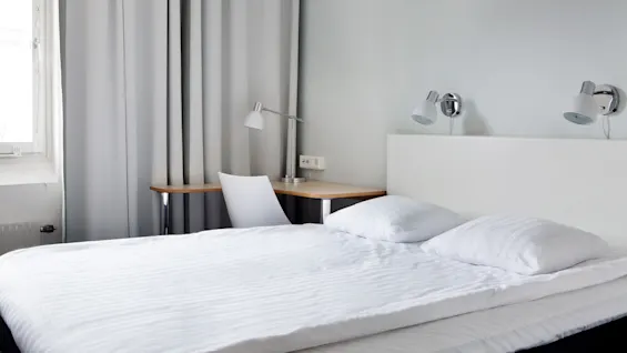 A neatly made bed with white linens in a minimalist room, adjacent to a desk with a lamp and chair, curtained window in the background.