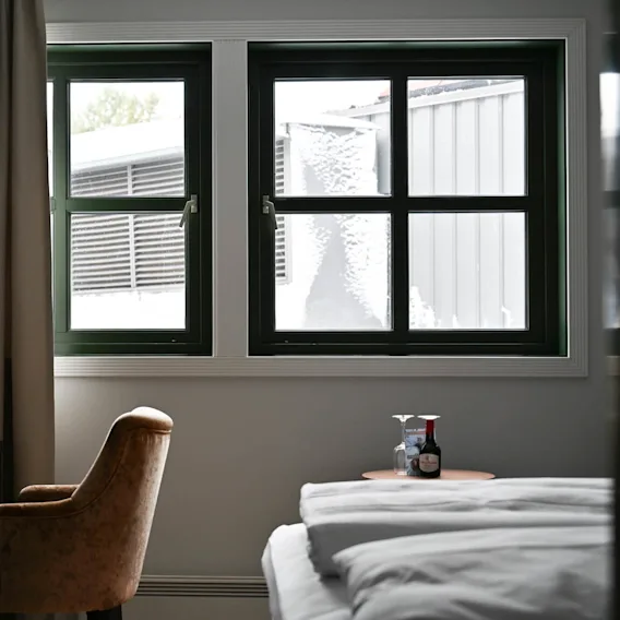 A cozy hotel room features a brown chair and desk by a curtained window overlooking a snowy scene. A small table beside the bed holds wine and glasses.