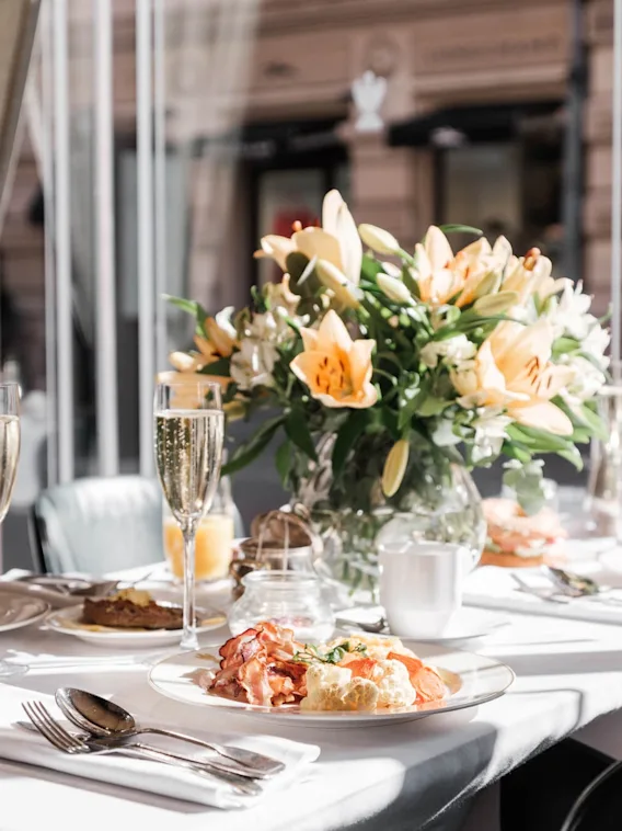 Et pent dekket frokostbord med champagne, bestikk og en fargerik blomsteroppsats i et solfylt rom med vinduer på Hotel Kämp i Helsinki.