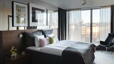 A modern hotel room with a comfortable bed, a stylish chair by a large window, and a city view.