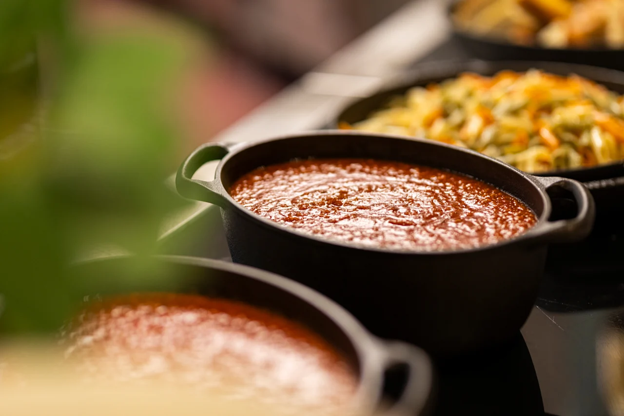 Close-up of a buffet line with a black pot of rich red sauce in the foreground and another pot of vegetables behind it.