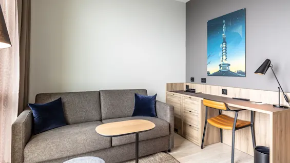 A gray sofa with blue cushions is set against a light wall. Adjacent is a wooden desk with a lamp and chair, featuring a geometric artwork above, in a modern room setting.