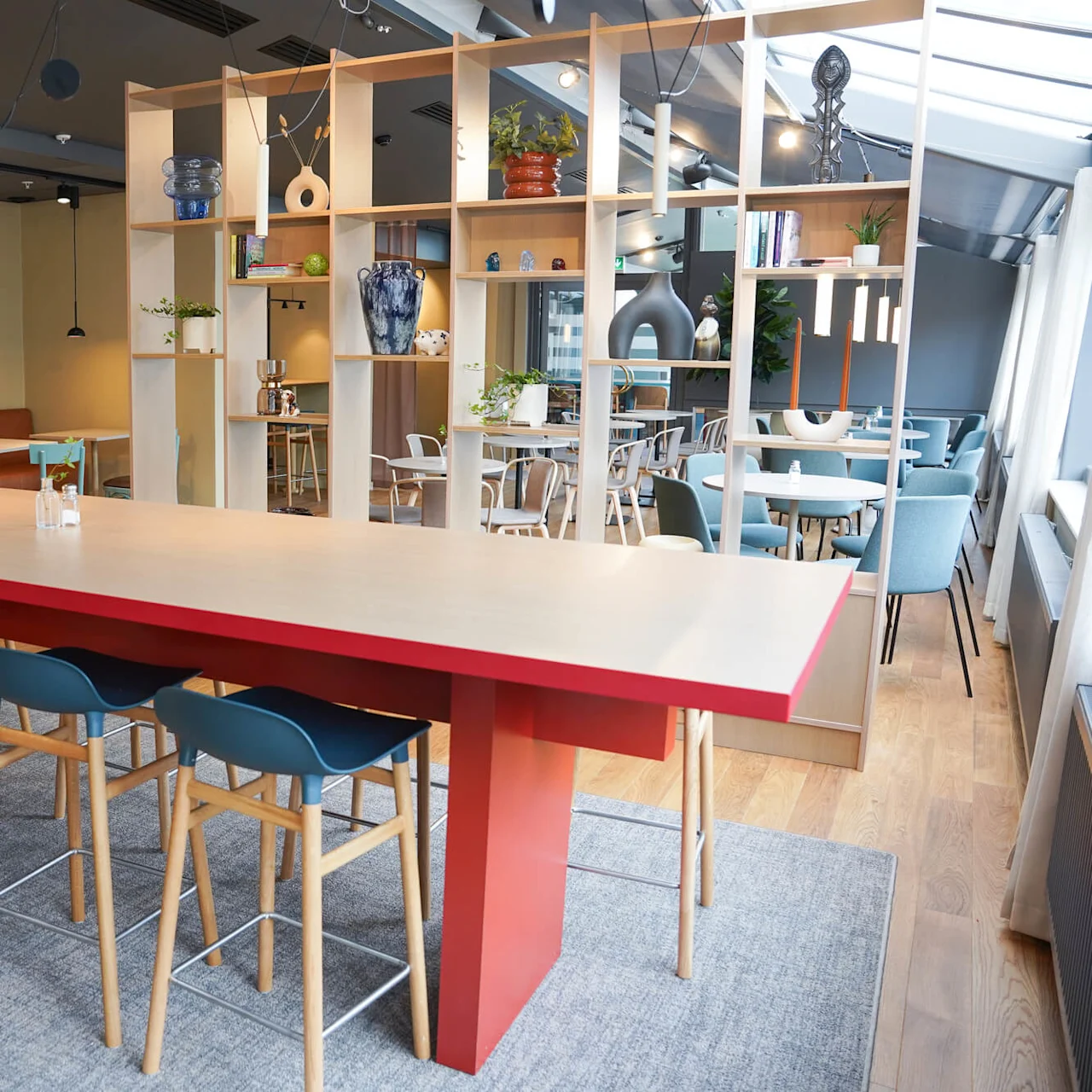 A long wooden table with blue chairs stands in a modern cafe, surrounded by shelves, plants, and additional seating areas.