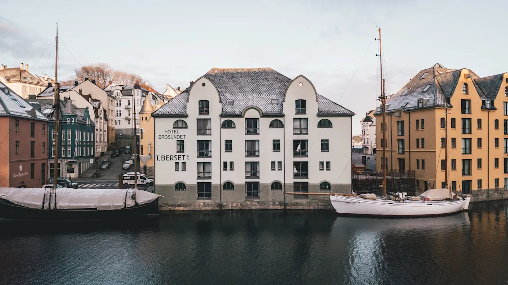 En hvid bygning mærket "Hotel Brosundet T. Berset AS" står ved en rolig vandkant, omgivet af farverige bygninger og fortøjede både under en klar himmel.