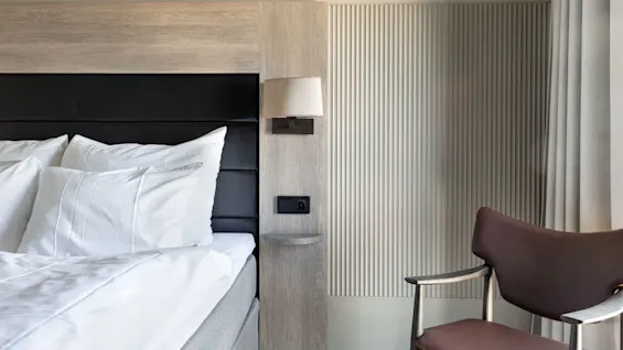A bed with white pillows and sheets sits beside a wooden headboard with an attached lamp, near a brown chair and beige curtains in a modern room.