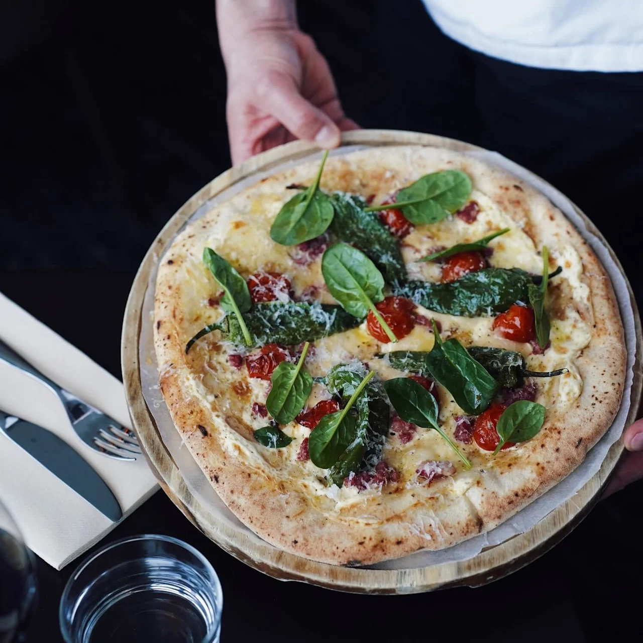 En pizza toppet med spinat, cherrytomater og ost holdes av en tatovert arm i hvit uniform. I nærheten er gaffel, kniv og glass.