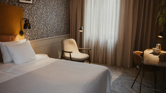 Mukava hotellihuone, jossa on sänky, viihtyisä nojatuoli ikkunan ääressä ja työpöytä lampun kanssa.