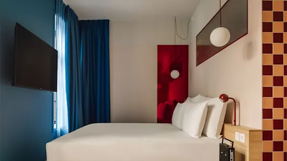A modern hotel room with a bed, blue curtains, a TV, and a colorful side table, featuring bold design elements.