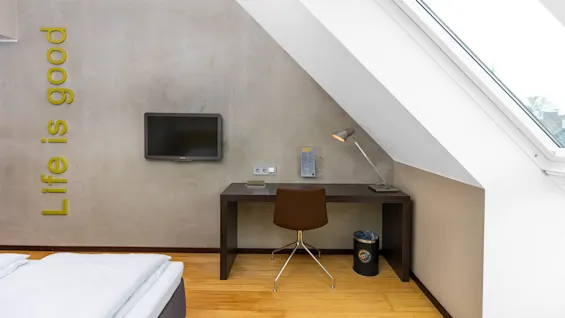 A desk with a chair and lamp sits beneath a slanted window in a room. A TV is mounted on a concrete wall with text: "Life is good."