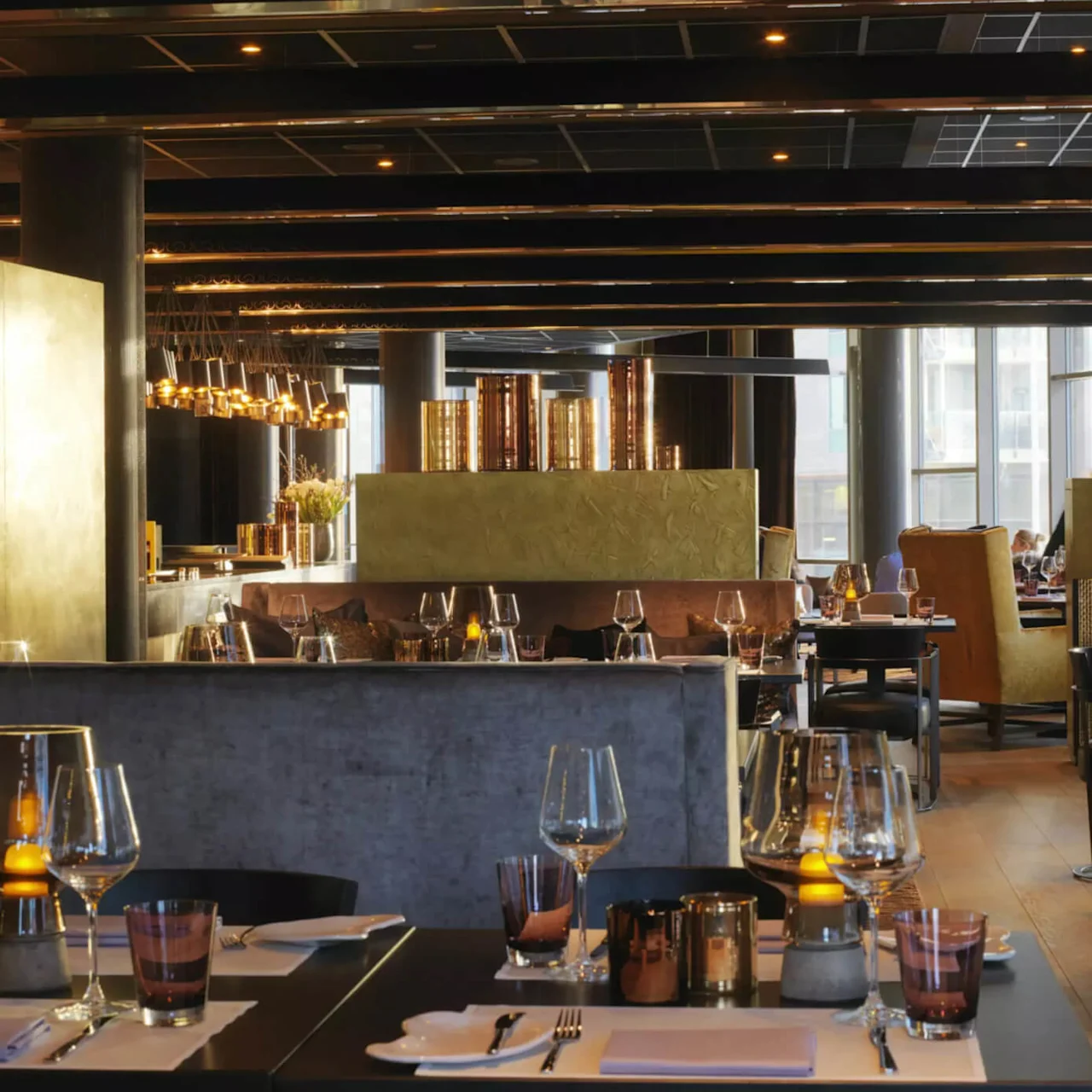 Restauranter borde arrangert elegant med vinglass og lysestaker, under moderne belysningsarmaturer i et sofistikert, svakt opplyst urbant miljø. Store vinduer gir naturlig lys i bakgrunnen.