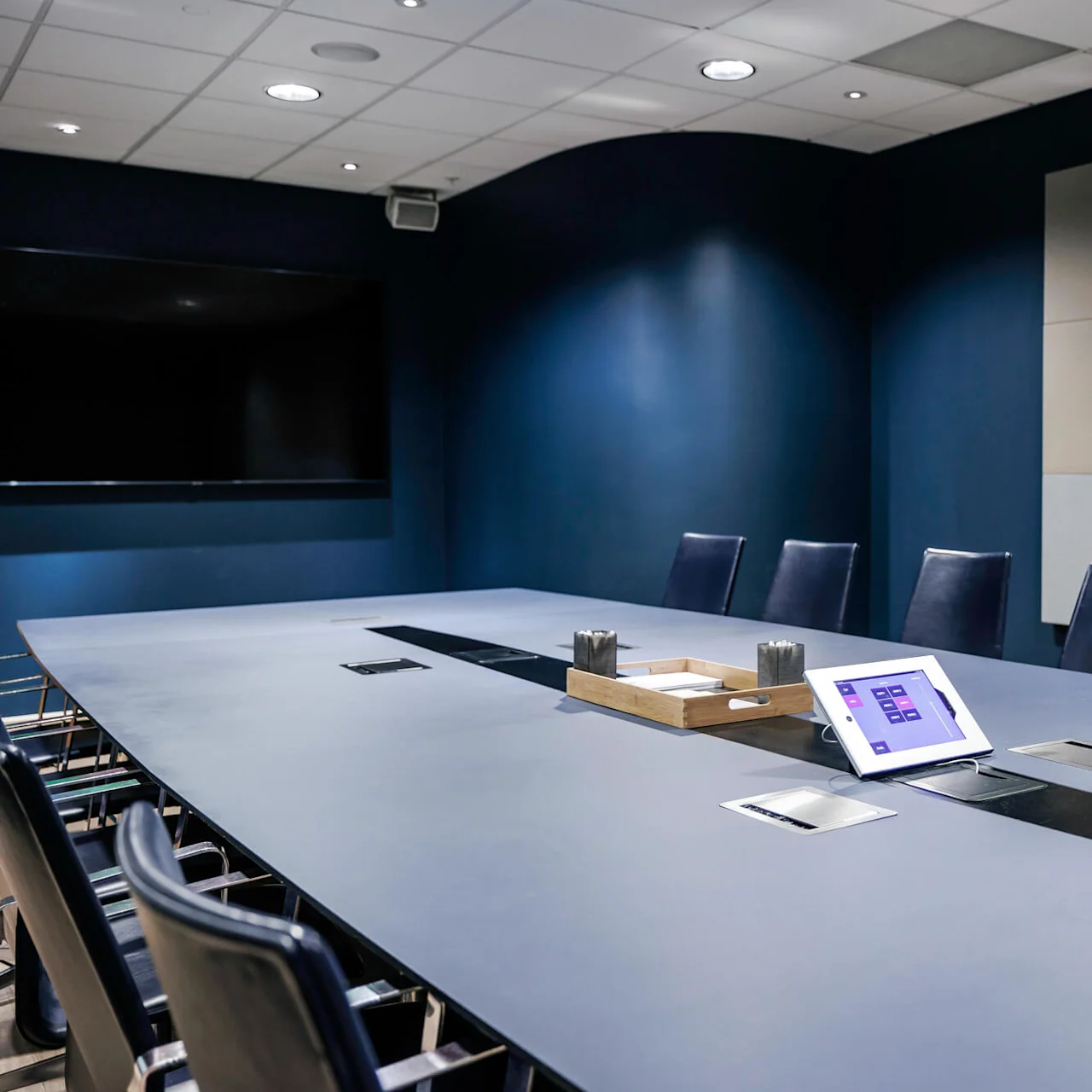A large conference table with chairs surrounding it, featuring a tablet and organizer tray on its surface, situated in a modern, dimly-lit meeting room.