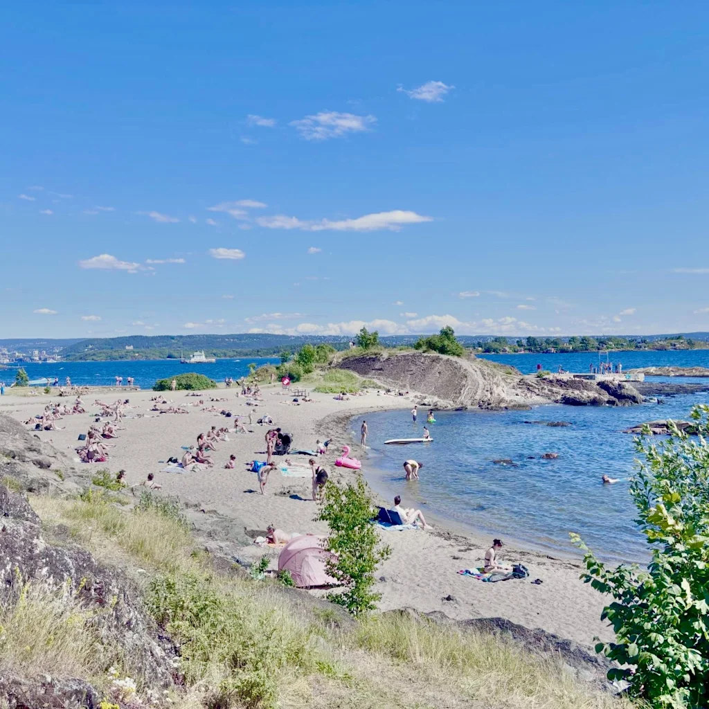 Människor kopplar av och simmar på en solig strand med kullar och blått vatten i bakgrunden. Huk i Oslo.