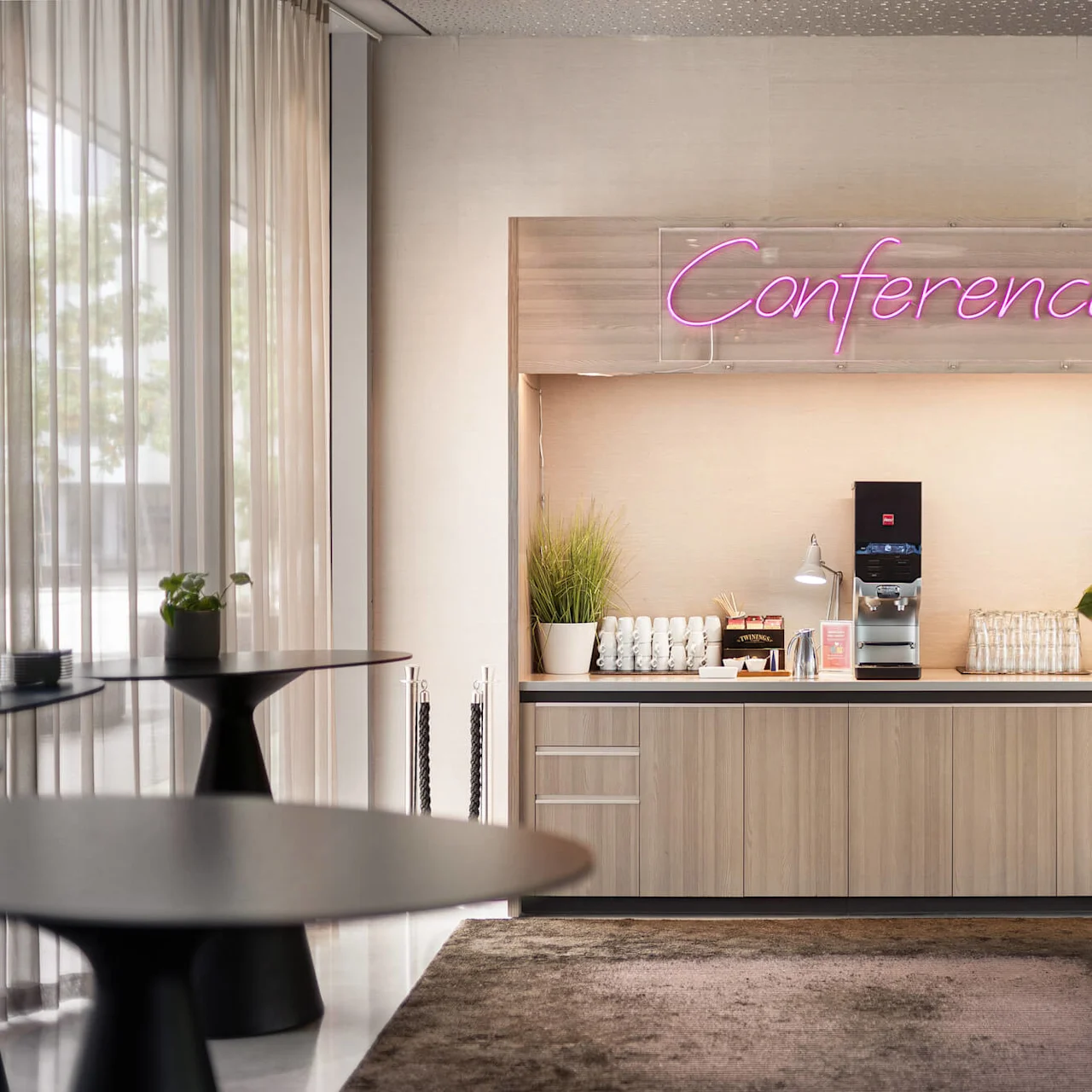 Coffee station with cups, plants, and a machine, situated under a neon "Conference" sign in a modern, well-lit room. A round table and curtains are nearby.