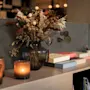 En hyggelig scene med en vase med tørrede blomster, tændte stearinlys og bøger på en hylde, der skaber en varm og indbydende atmosfære.