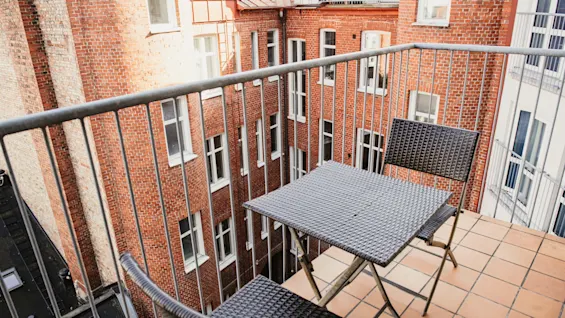 En hyggelig balkon med bord og stole, der tilbyder udsigt over en solbeskinnet gårdhave med murstensbygninger.