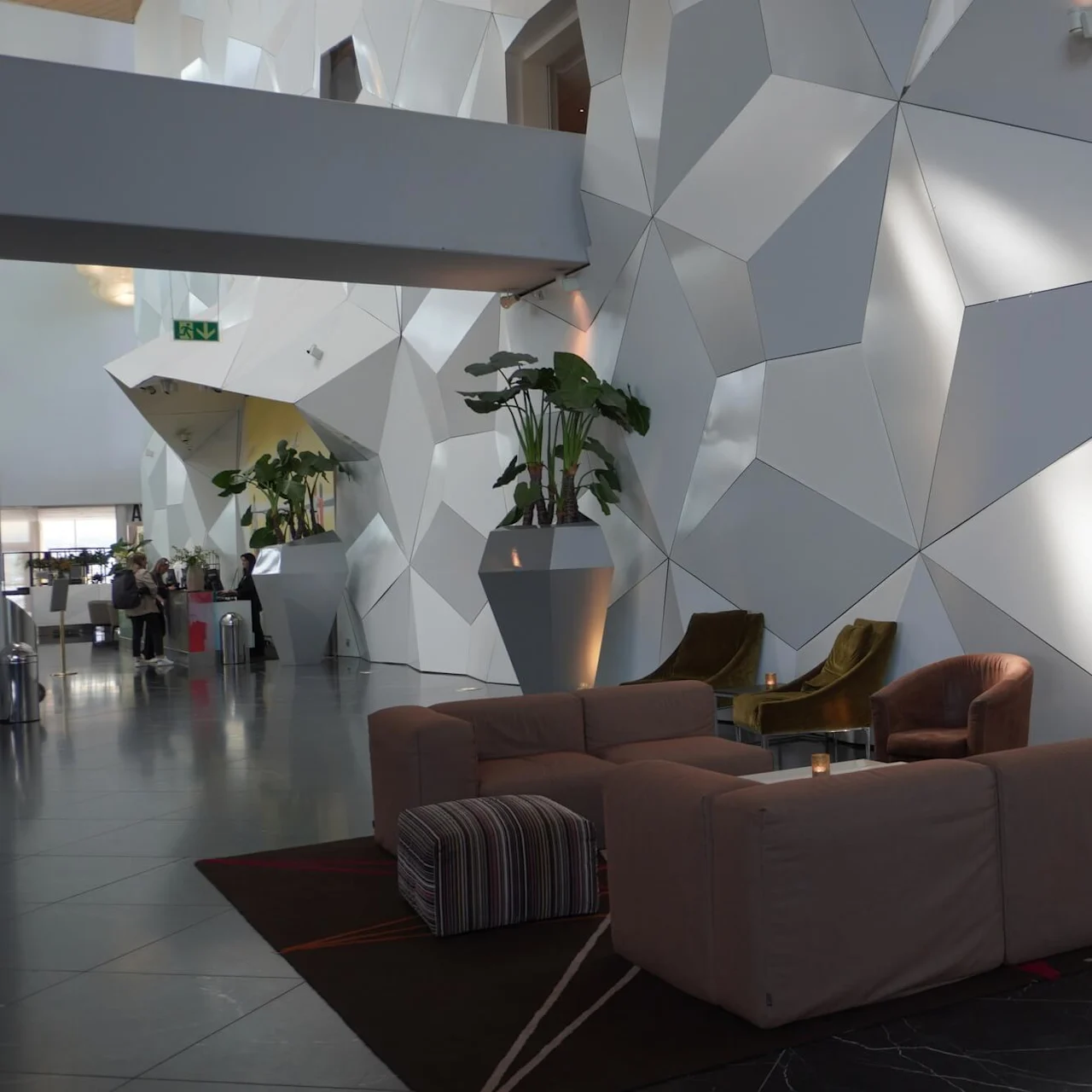 Modern lounge area features geometric wall panels, comfortable sofas, and armchairs. Large potted plants enhance the contemporary decor, while people sit at a distant bar. Green exit sign visible.