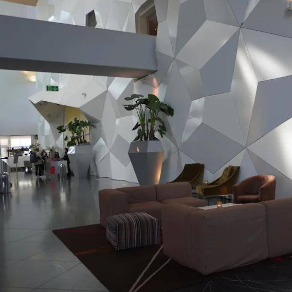Moderne loungeområde med geometriske vægpaneler, komfortable sofaer og lænestole. Store potteplanter fremhæver den moderne indretning, mens folk sidder ved en fjern bar. Grøn udgangsskilt synlig.