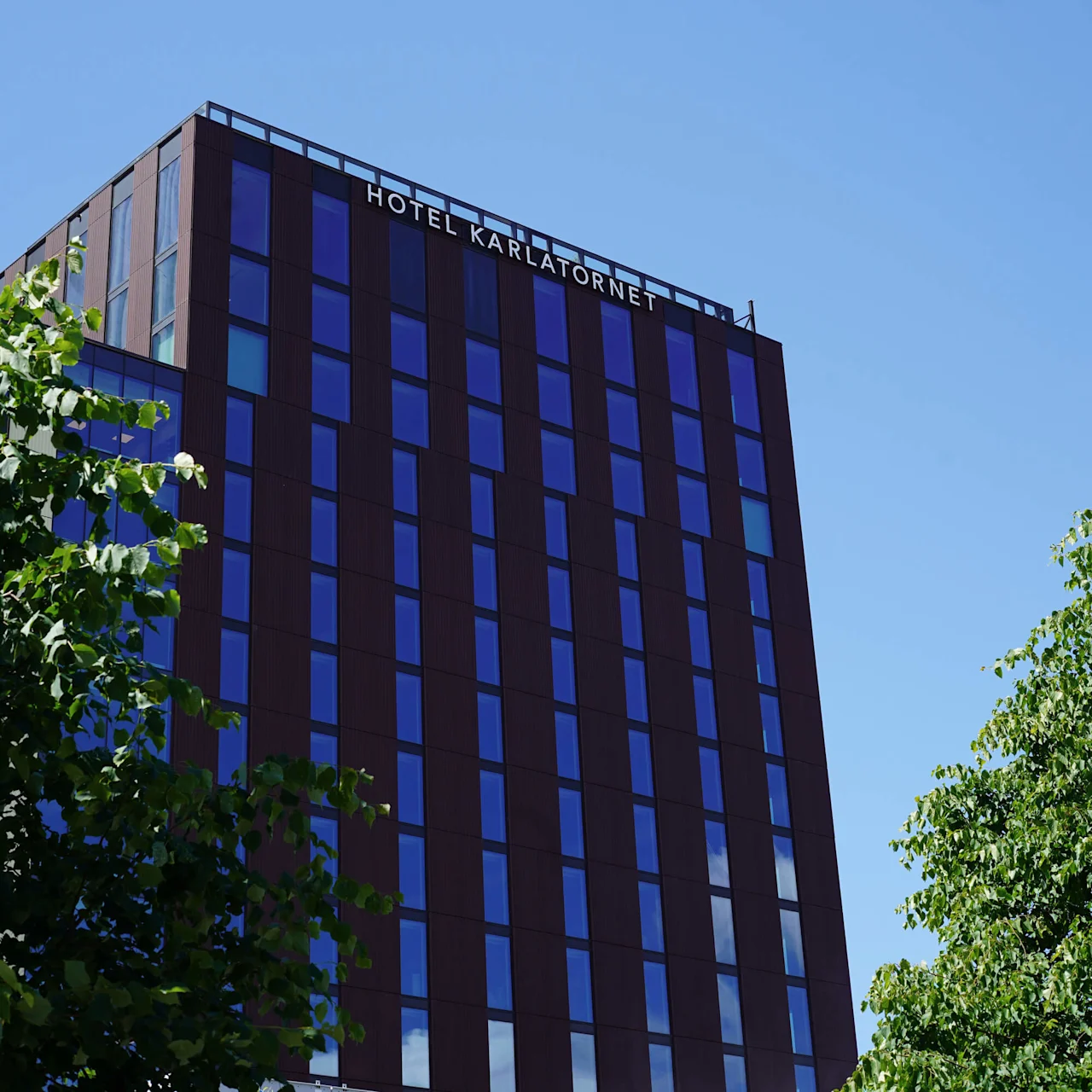 En hög byggnad märkt "HOTEL KARLATORNET" med reflekterande glasfönster står mot en klarblå himmel. Gröna träd ramar in förgrunden.