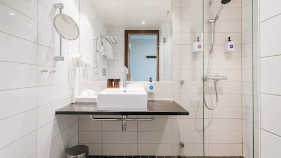 Ett ljust och modernt hotellbadrum med vitt kakel, handfat på en mörk bänkskiva och en duschhörna med glasvägg.