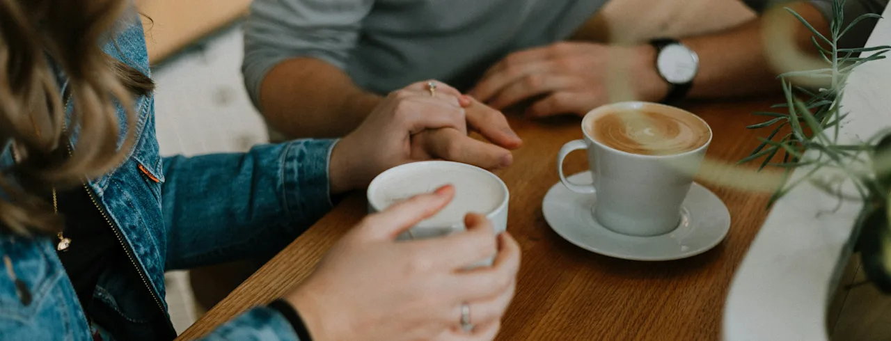 Två personer delar en lugn stund över kaffe, händer vilar lätt på ett träbord, njuter av sina drycker.