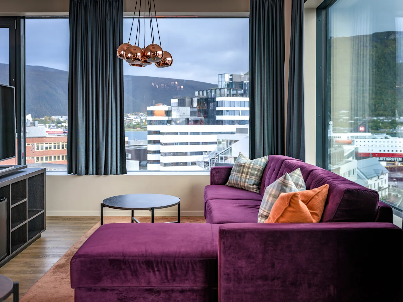 Tyylikäs olohuone, jossa on violetti kulmasohva, moderni valaistus ja panoraamanäkymä kaupunkiin ja vuorille.