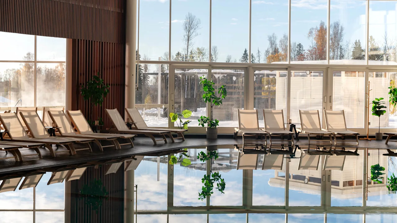 Poolstole er arrangeret på række ved en stor indendørs pool, der reflekterer udsigten over træer og himmel gennem store vinduer. Potteplanter dekorerer rummet.